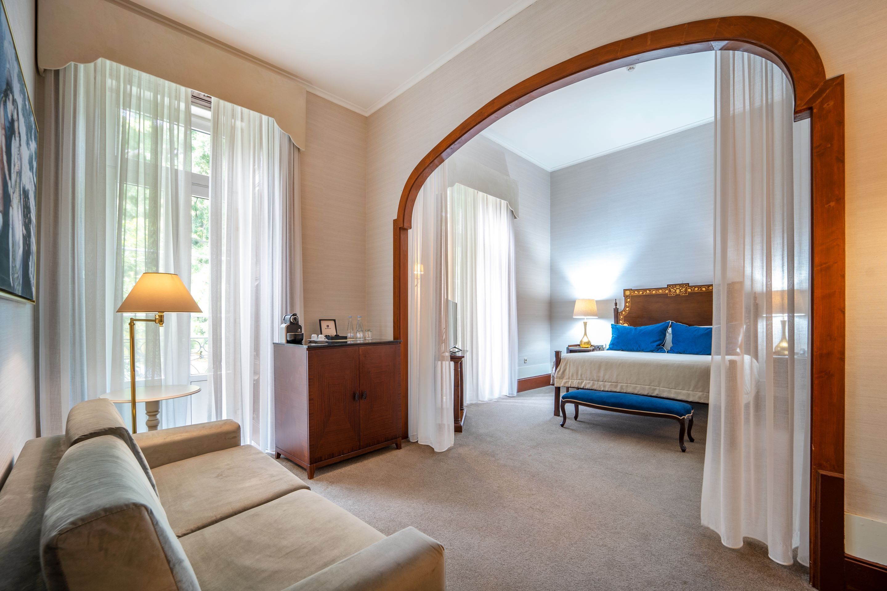 A hotel room with a sofa, a bed with blue pillows, a wooden headboard, a desk with a lamp, and a view through an arched doorway.