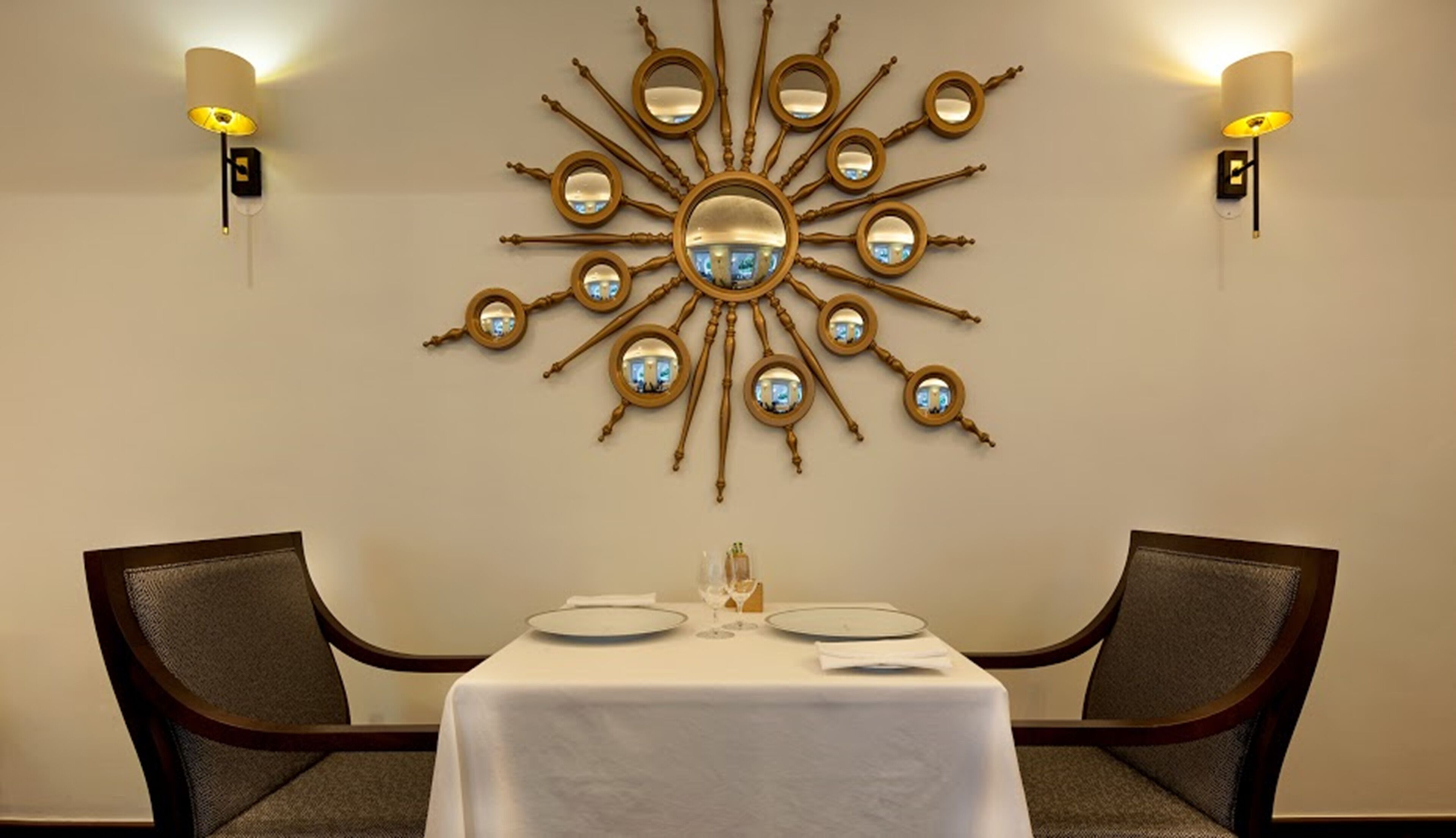 A dining area with a central round mirror surrounded by smaller mirrors, two chairs, a table with a white tablecloth, and wall-mounted lamps.