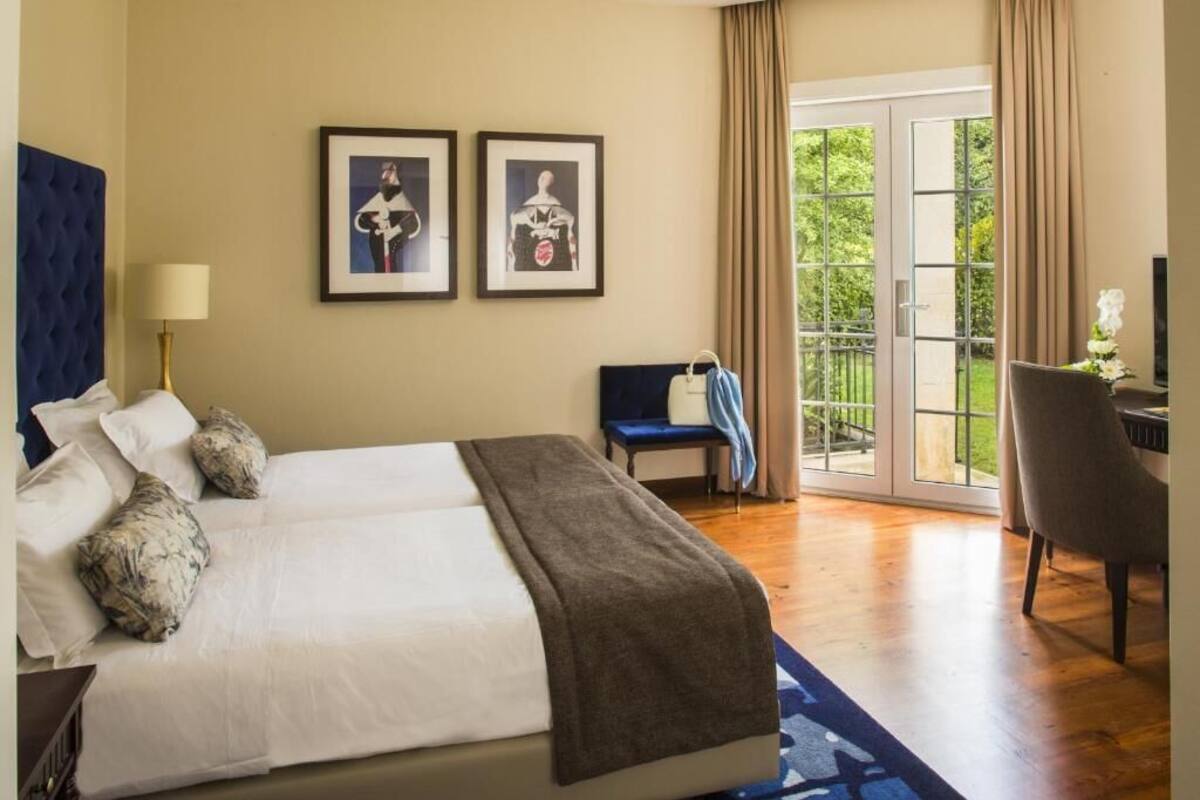 Une chambre d’hôtel avec un grand lit, un bureau et une chaise. Deux tableaux encadrés ornent le mur, ainsi qu’une lampe. Une porte coulissante offre une vue sur l’extérieur.