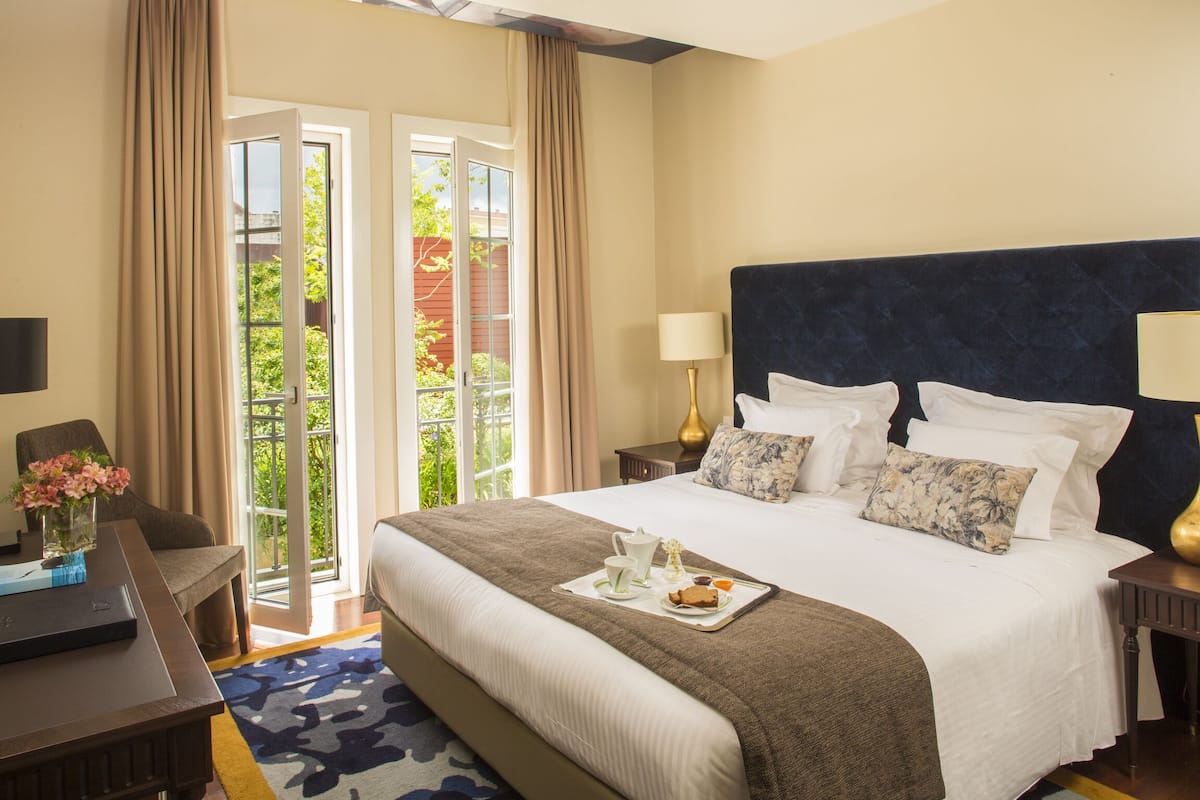 Une chambre d’hôtel avec un grand lit, un bureau, une chaise et une vue sur un jardin, accessible par une porte vitrée.