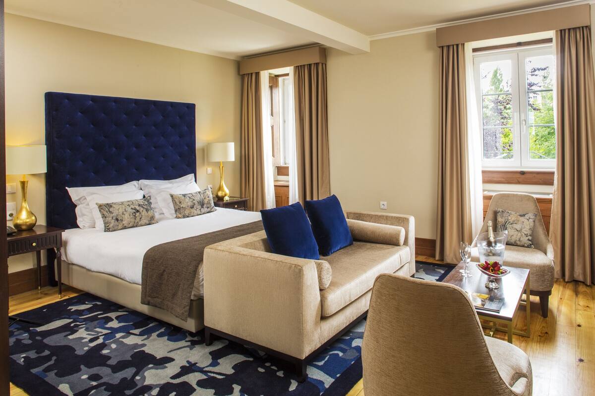 Une chambre d’hôtel avec un grand lit, un canapé, un fauteuil, une petite table avec un verre d’eau et une bouteille, et une vue sur la verdure par la fenêtre.