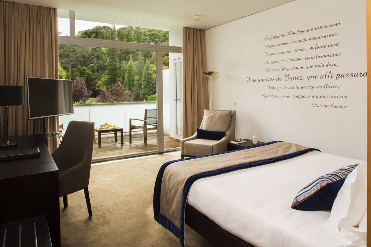 Une chambre d’hôtel comprenant un lit, un bureau, une chaise, une télévision et un balcon avec vue sur les arbres.