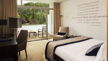 Habitación de hotel con cama, escritorio, silla, televisión y balcón con vistas a árboles.