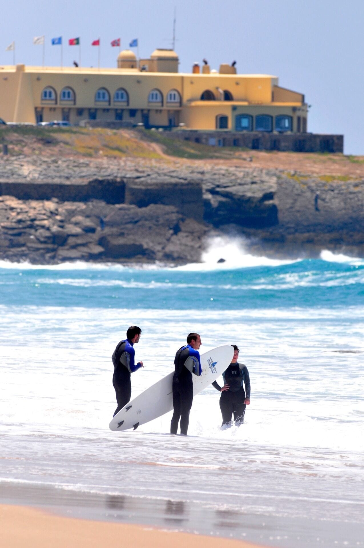Un edificio con varias banderas en el techo, cerca de una playa donde las olas rompen sobre la orilla.