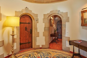 A hallway with stone arches, a wooden door, a lamp, a table, and a framed picture on the wall.
