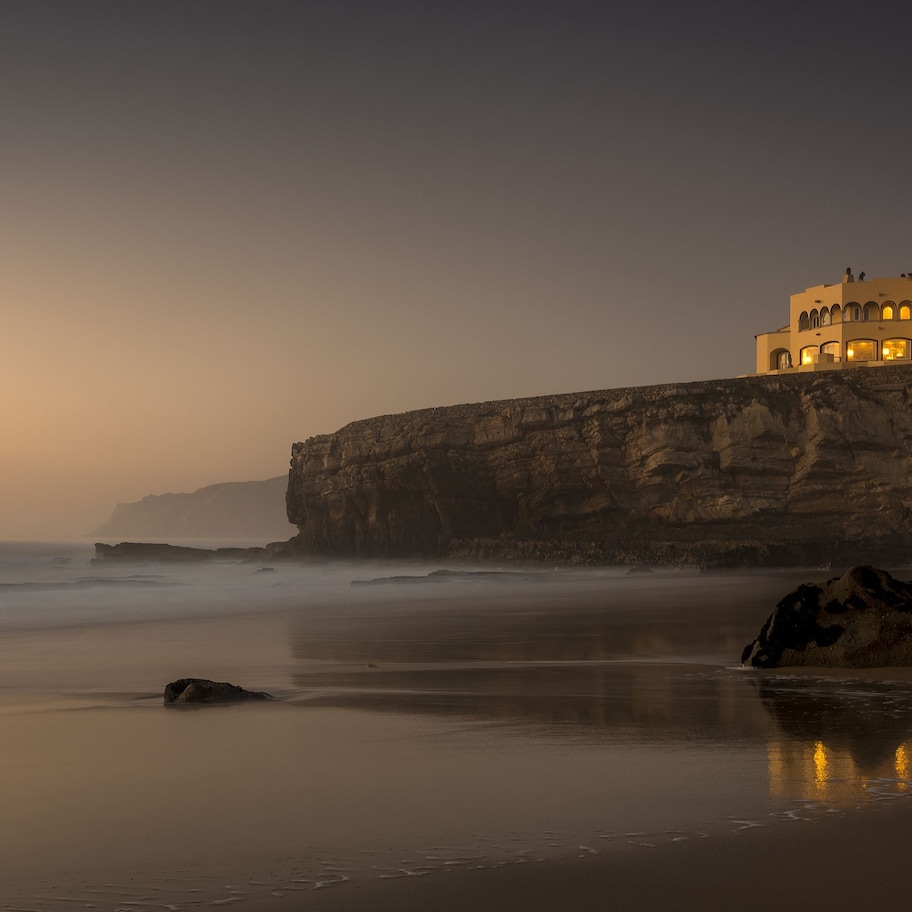 A coastal hotel with a cliffside view, calm beach, and ocean waves.