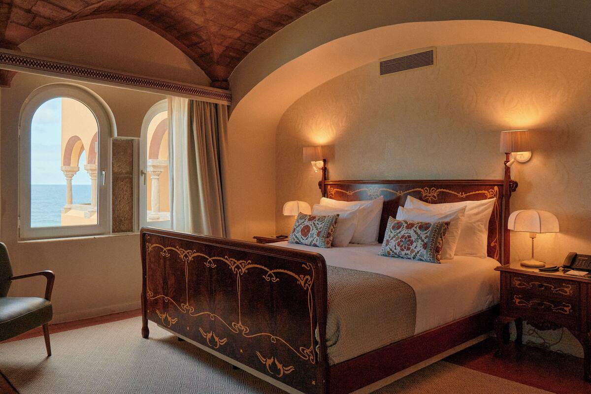 Un dormitorio con una cama grande, dos lámparas de noche, una silla y vistas al mar a través de ventanas de arco.