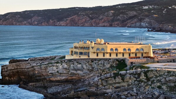 Un edificio costero con techo plano y múltiples ventanas, ubicado en un acantilado rocoso con vistas al mar.