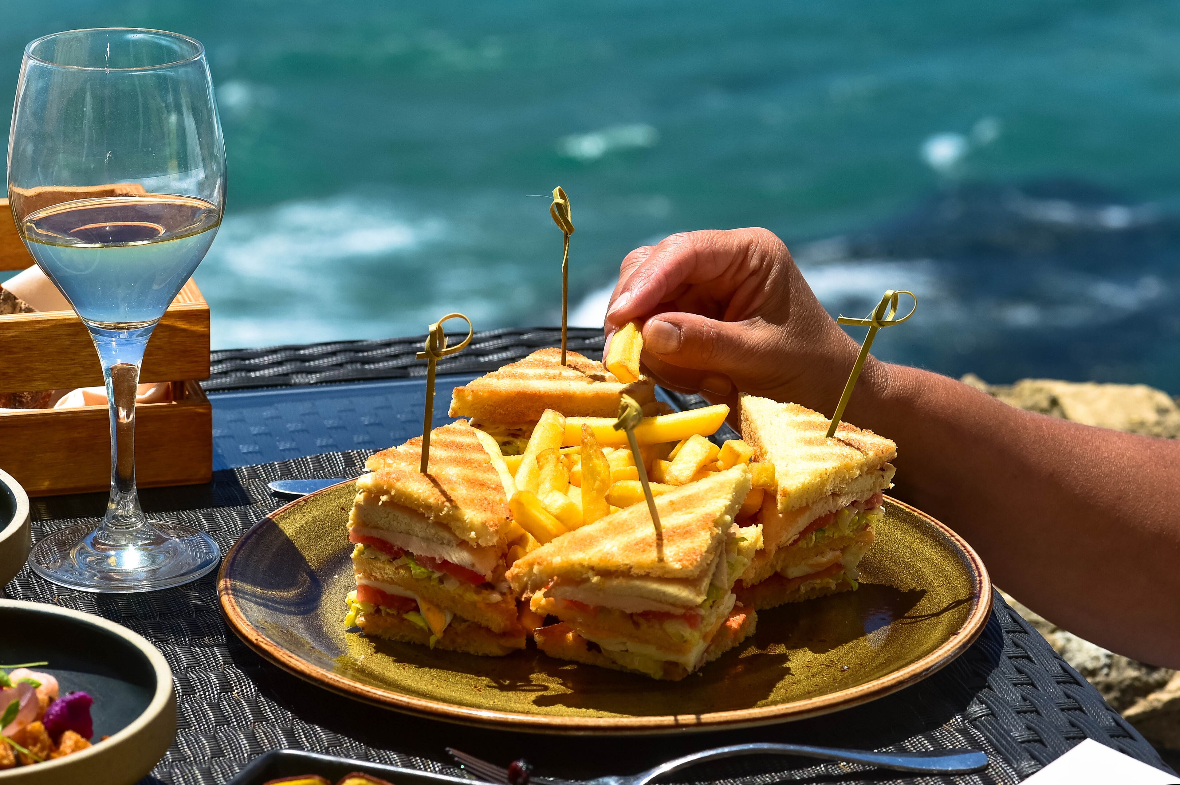 Un sándwich con palillos de dientes sobre un plato, una copa de vino y vista al océano.