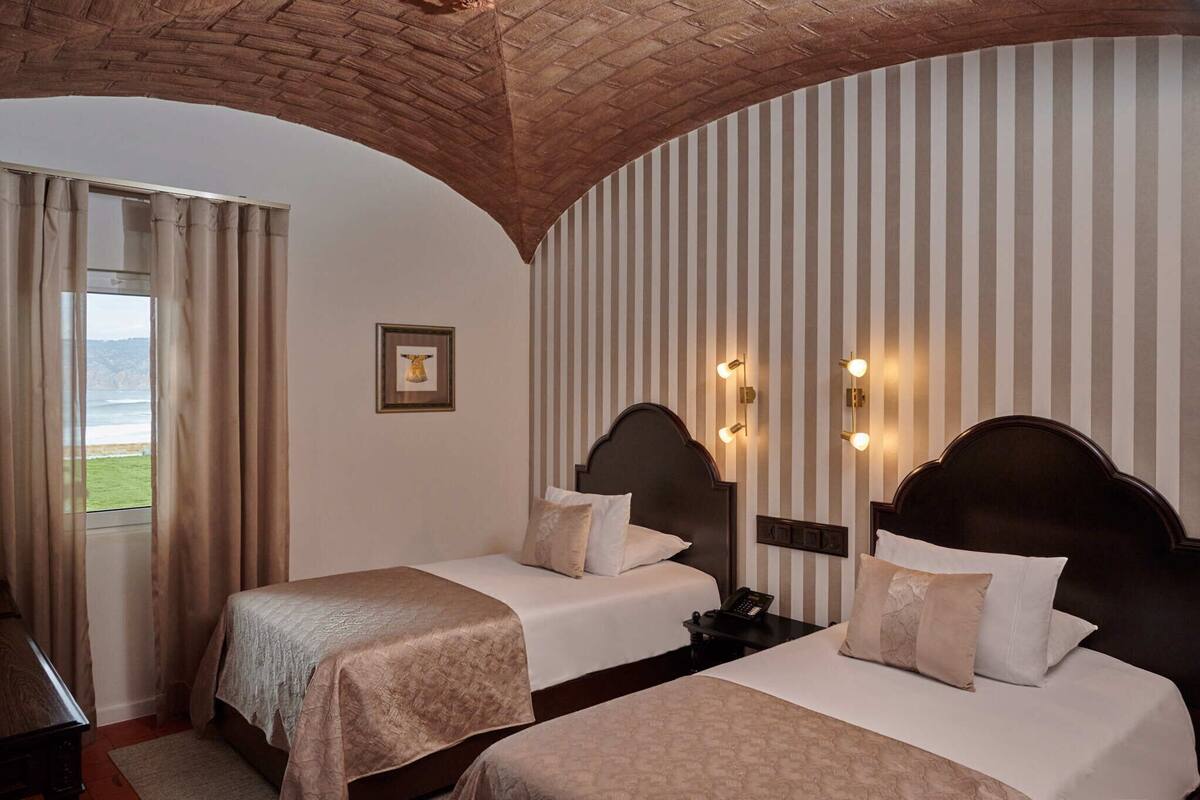 Habitación de hotel con dos camas, vista al mar, papel tapiz a rayas y techo de ladrillo.