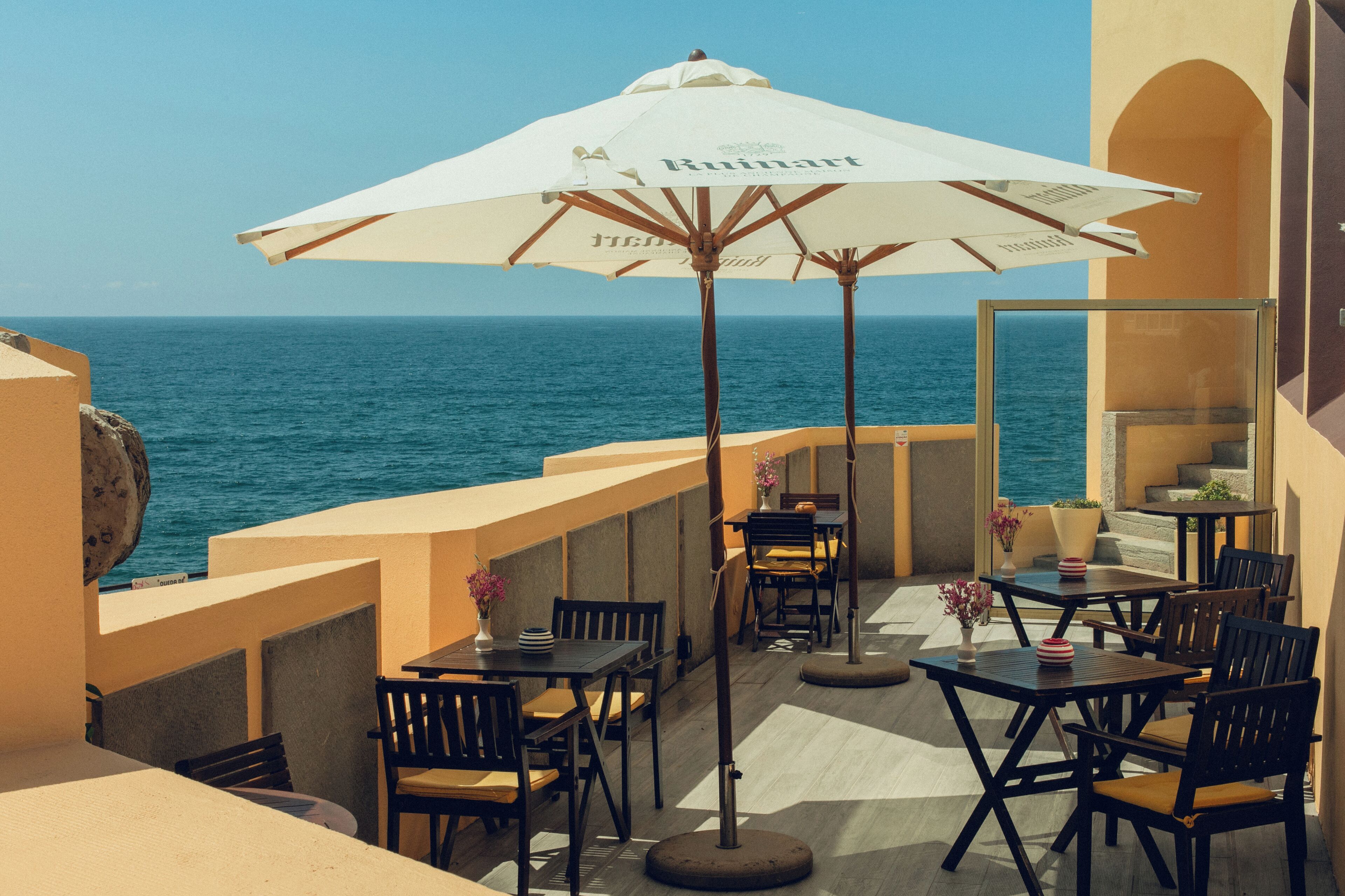 Una terraza con mesas y sillas, un paraguas grande y vista al mar.