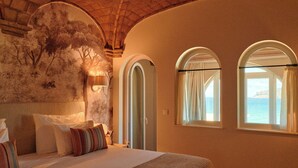 Ein Schlafzimmer mit einem großen Bett, zwei Bogenfenstern, einem Wandgemälde und Meerblick.