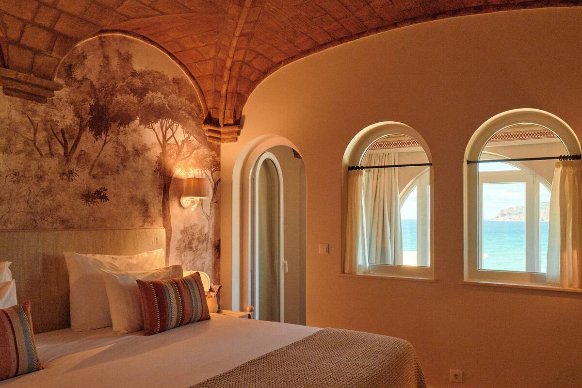 Un dormitorio con una cama grande, dos ventanas arqueadas, un mural en la pared y vistas al mar.