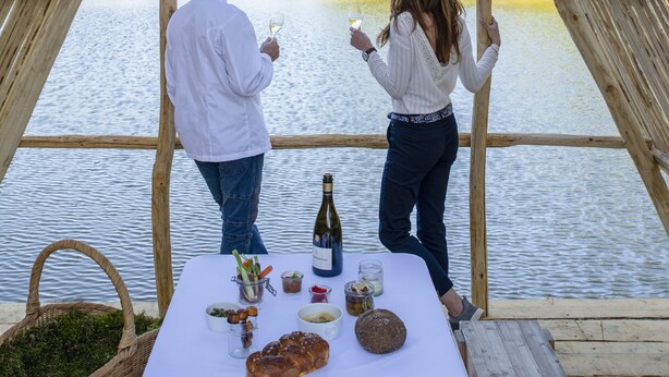 A wooden pergola structure overlooking a calm lake with a table set for a meal and a bottle of wine.