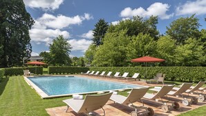 A poolside area with lounge chairs and umbrellas, surrounded by well-manicured lawns and trees.