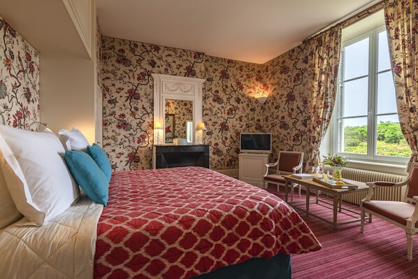 A hotel room with a large bed, a desk, a chair, a television, and a window with a view of greenery.