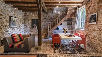 Ein rustikales Interieur mit einer Steinwand, Holzbalken, einem Sofa, einem Esstisch und einer Treppe.
