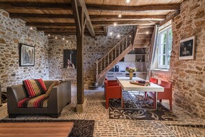 A rustic interior with a stone wall, wooden beams, a sofa, a dining table, and a staircase.