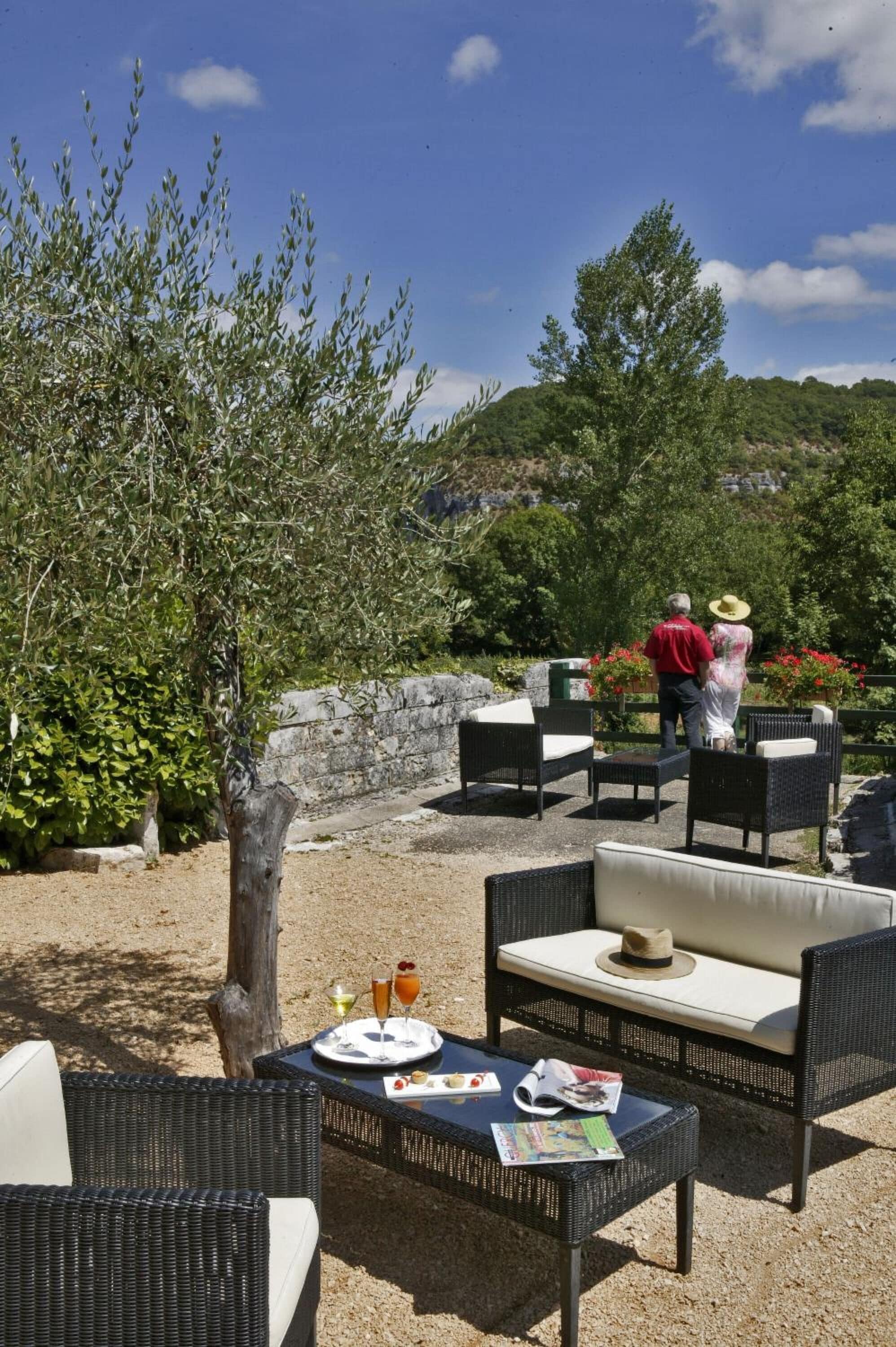 Un espace extérieur aménagé avec des meubles en osier et une table pour deux, donnant sur un jardin agrémenté d’un mur en pierre et d’arbres.