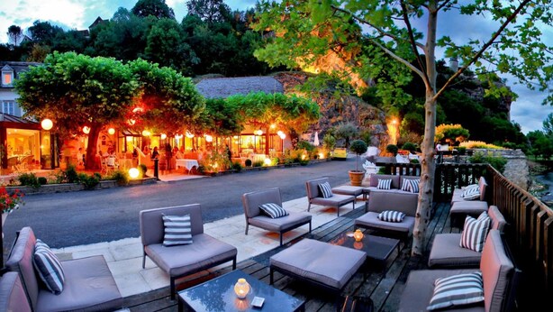 An outdoor seating area with cushioned chairs and tables, a restaurant in the background, and a view of a rocky hillside.