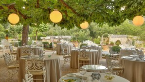 An outdoor dining area with round tables and white chairs, set against a backdrop of greenery and stone structures.