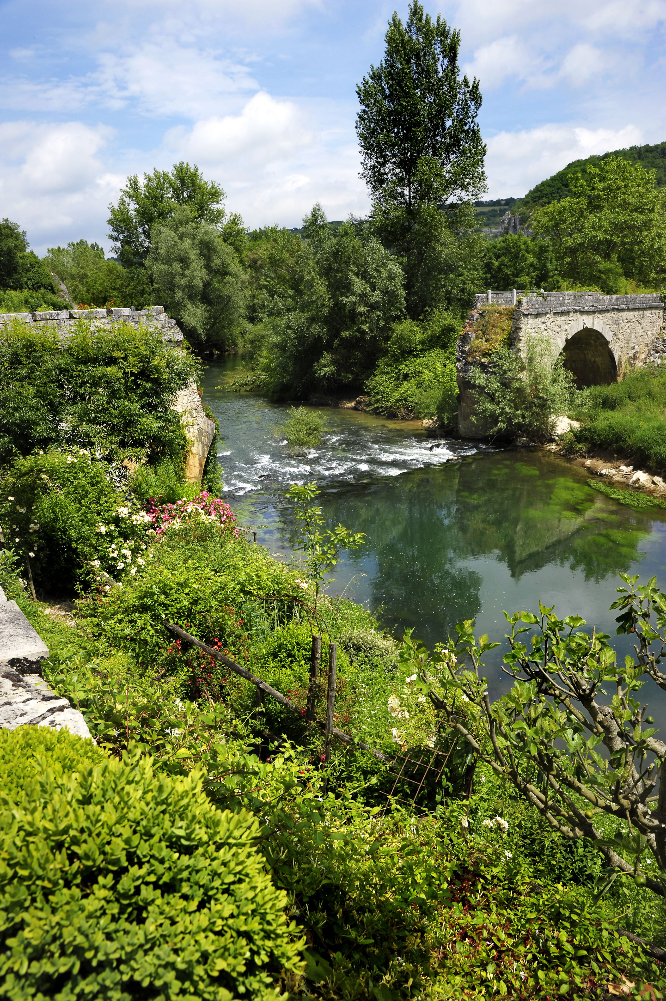A river flowing through a lush, green landscape with a stone bridge and a stone archway.