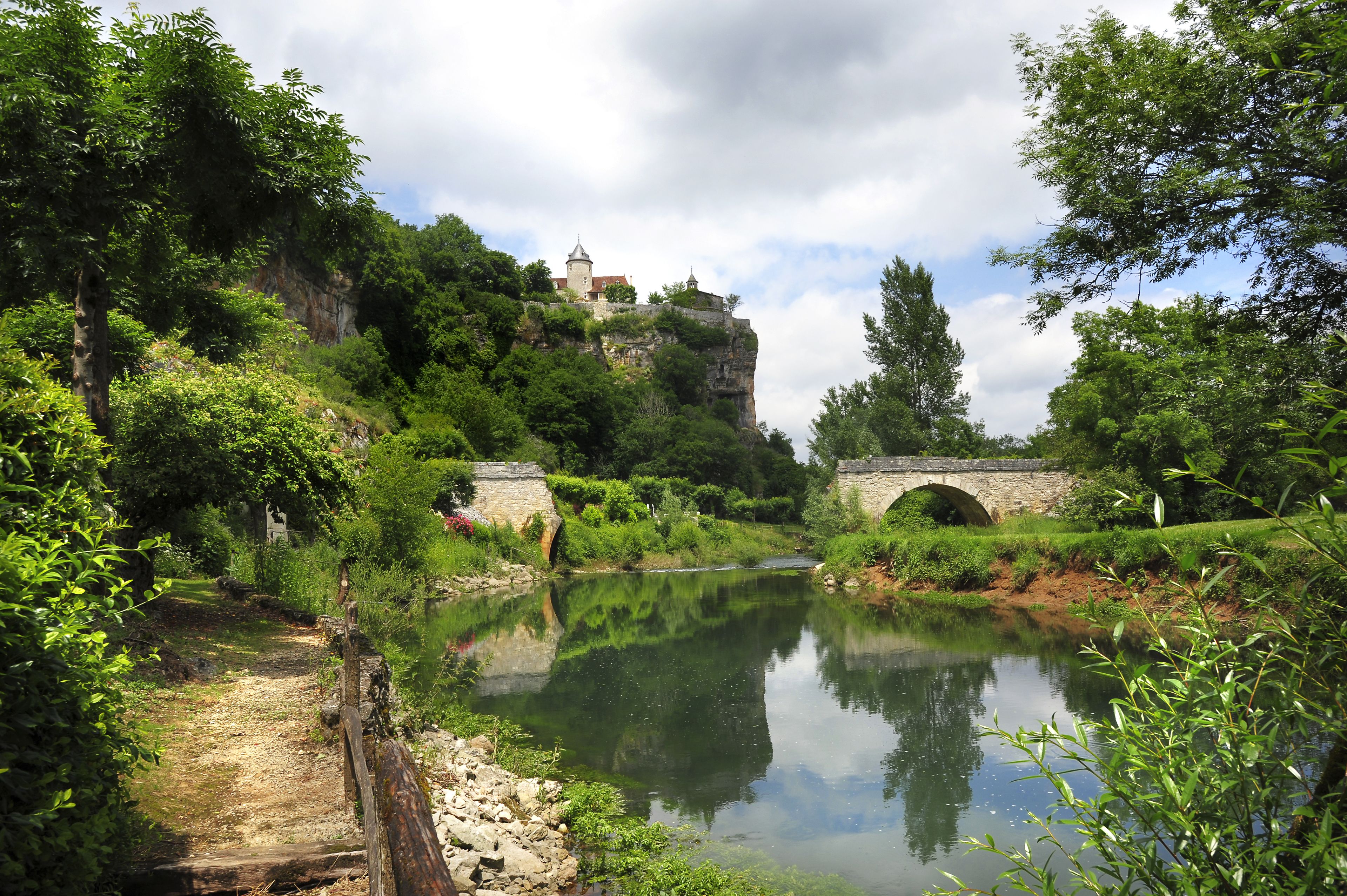 A serene riverside scene with a stone bridge, lush greenery, and a castle on a cliff.