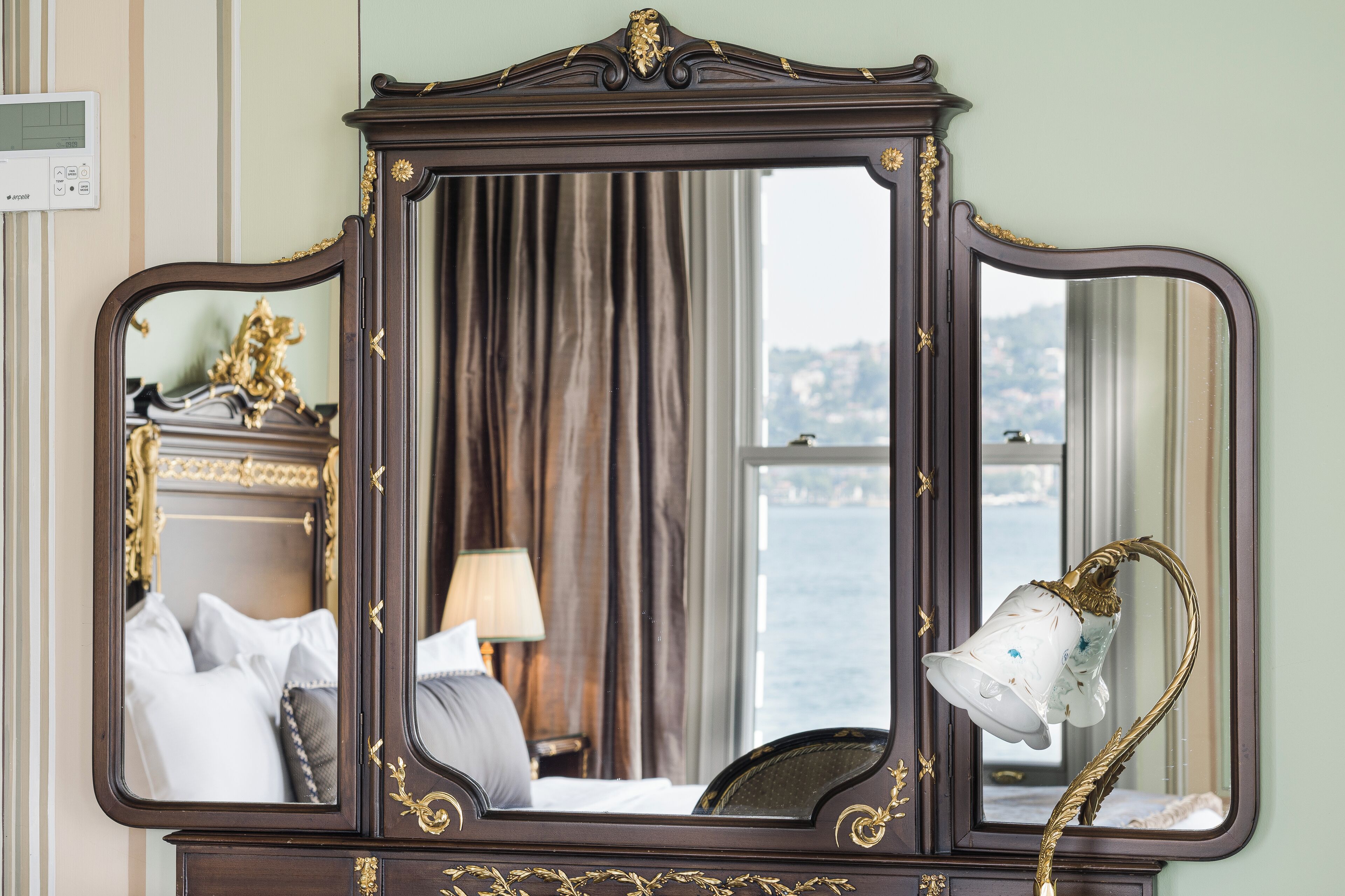 Un espejo con detalles ornamentales dorados, una cama con ropa blanca, una lámpara y vistas de una masa de agua a través de una ventana.