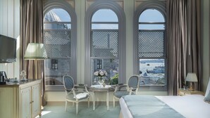 A hotel room with a bed, two chairs, a small table, a TV, and a view of a cityscape through large arched windows. - Bosphorus Palace Hotel - Special Class (Istanbul)