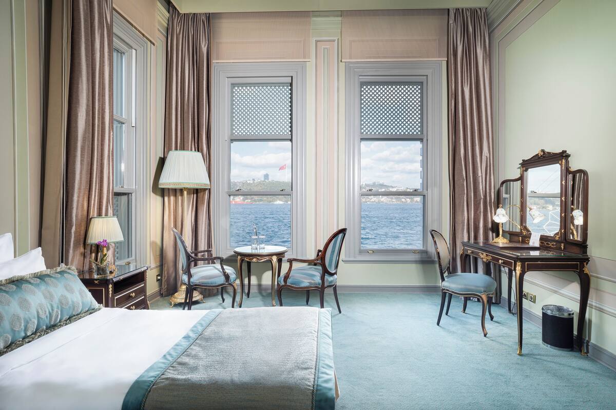 A hotel room with a bed, a desk with a mirror, two chairs, a lamp, and a view of the sea through large windows.