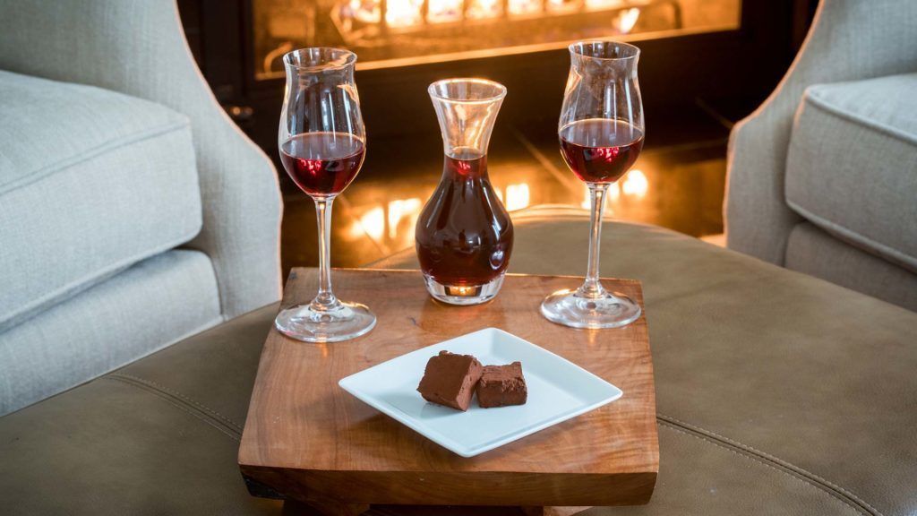 Un coin salon confortable avec une table en bois sur laquelle se trouve une assiette blanche contenant des chocolats et deux verres de vin rouge.