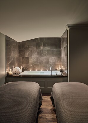 A modern bathroom with a large bathtub, two beds with matching bedding, and a stone wall.