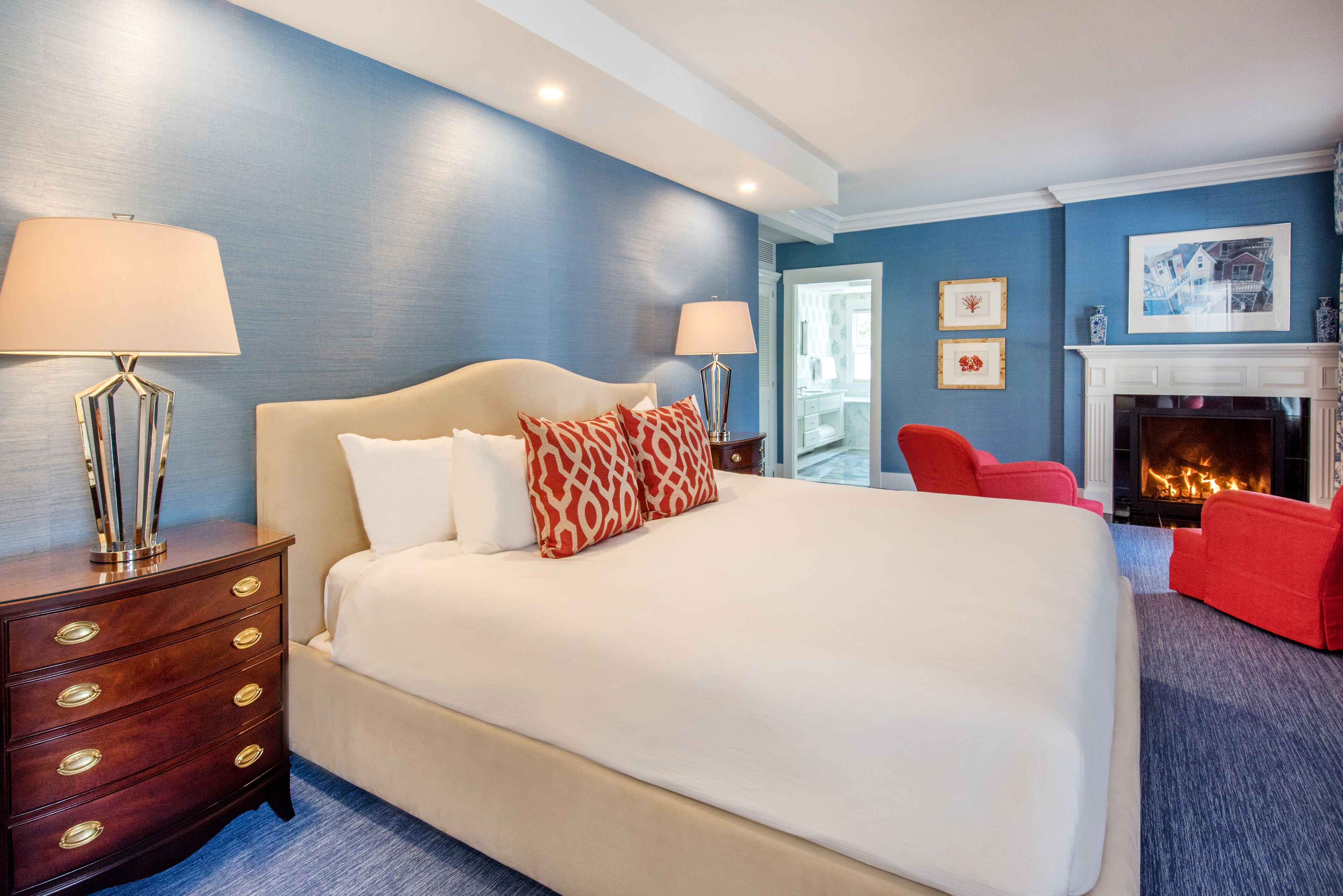 A hotel room with a large bed, two bedside lamps, a dresser with drawers, a red chair, a fireplace, and framed pictures on the wall.