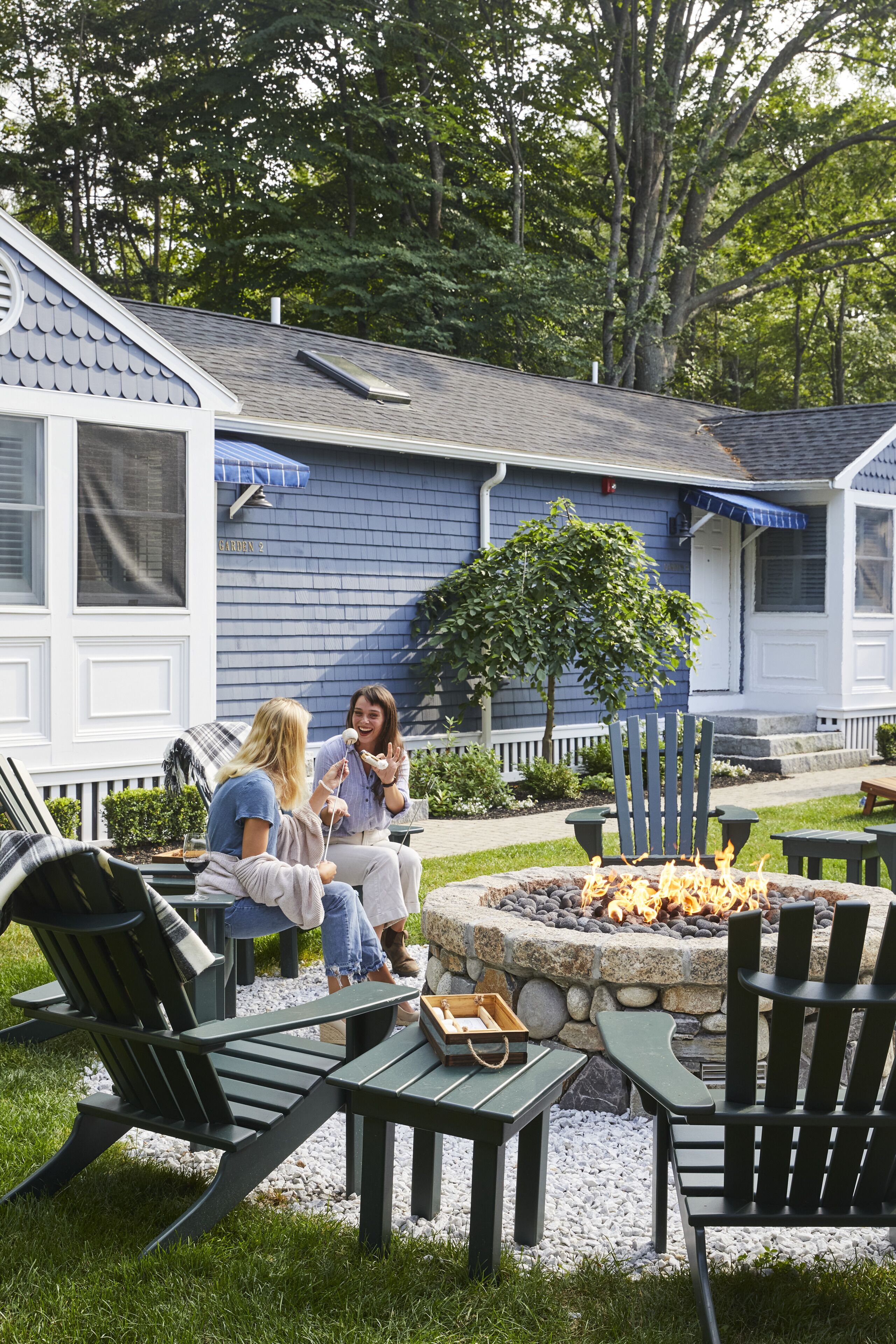 A cozy outdoor seating area with Adirondack chairs and a stone fire pit, surrounded by a blue building with a white porch.