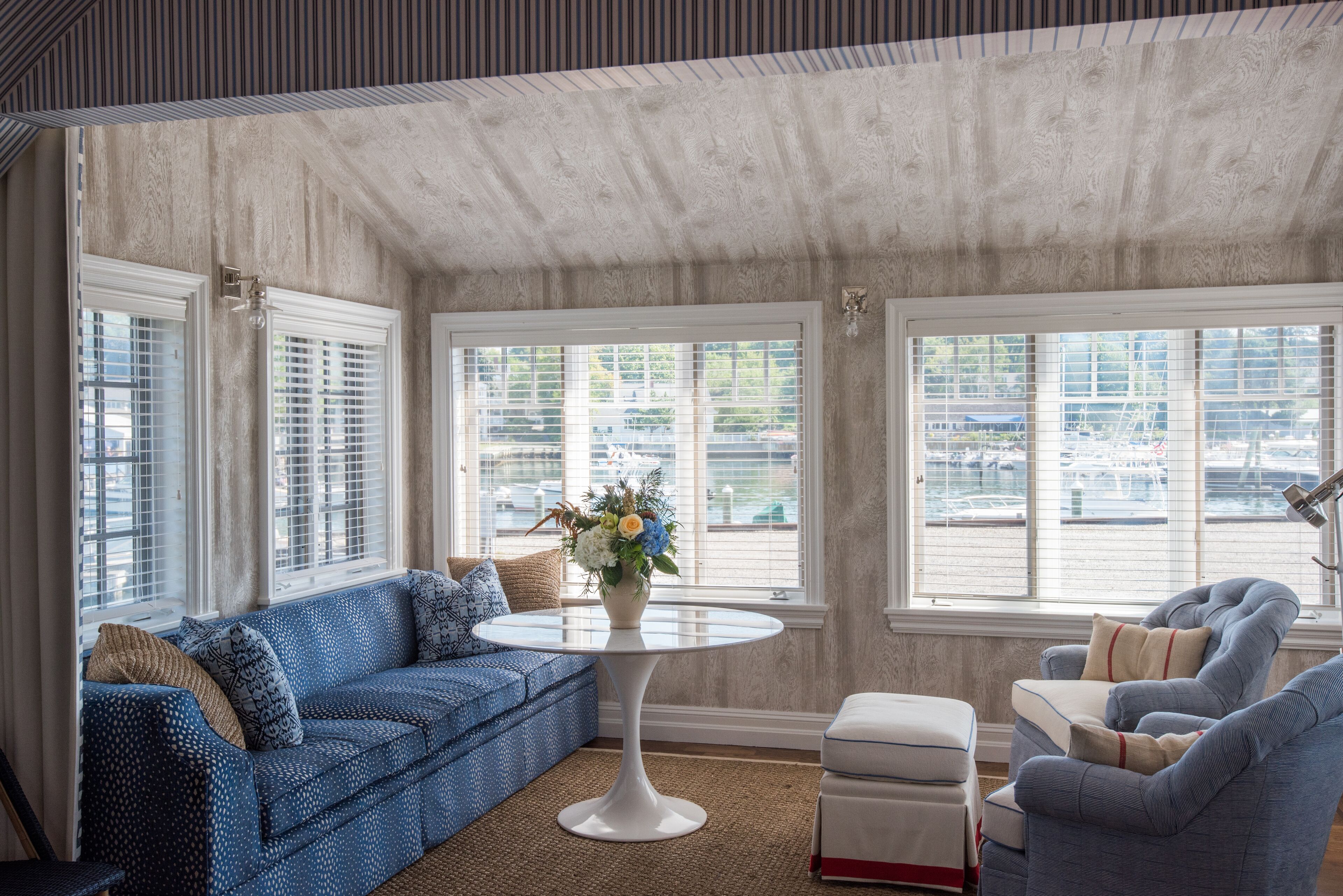 Una habitación con un sofá azul estampado, una mesa redonda blanca con un jarrón de flores y un sillón azul.