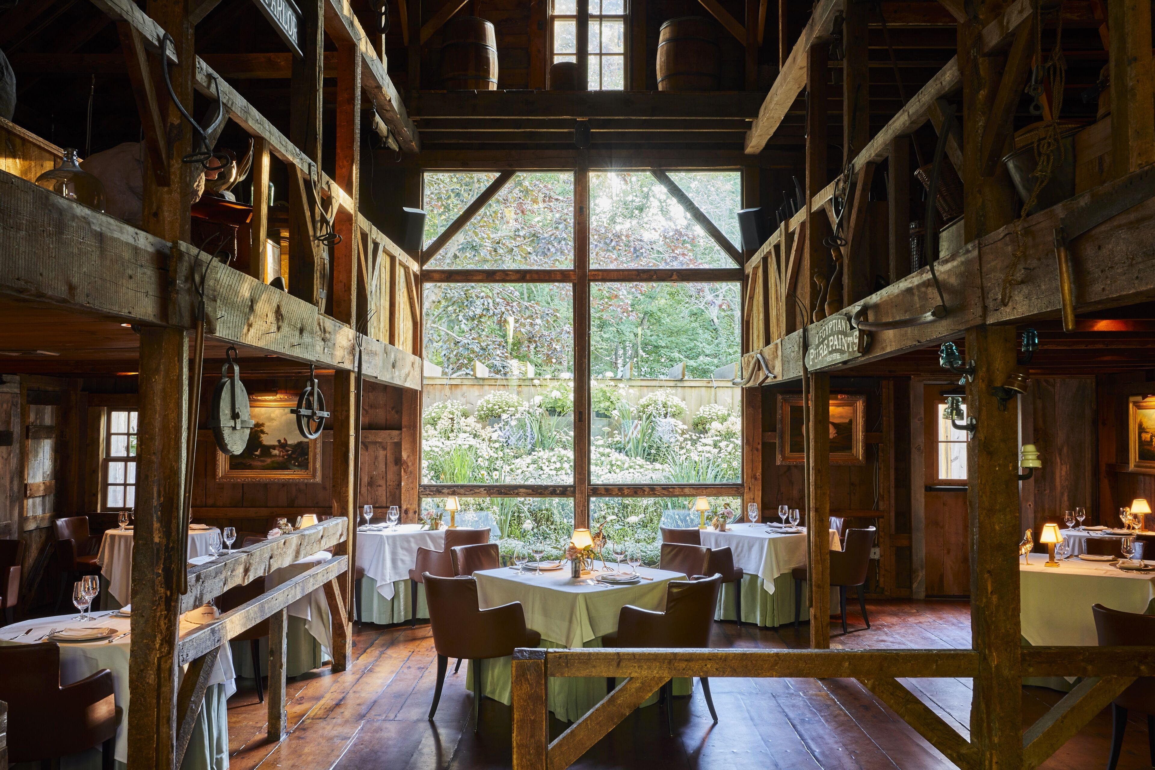 A rustic wooden dining area with tables and chairs, large windows, and a view of greenery outside.
