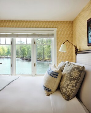 A bedroom with a large window overlooking a body of water and trees. There is a bed with two pillows and a bedside table with a lamp.