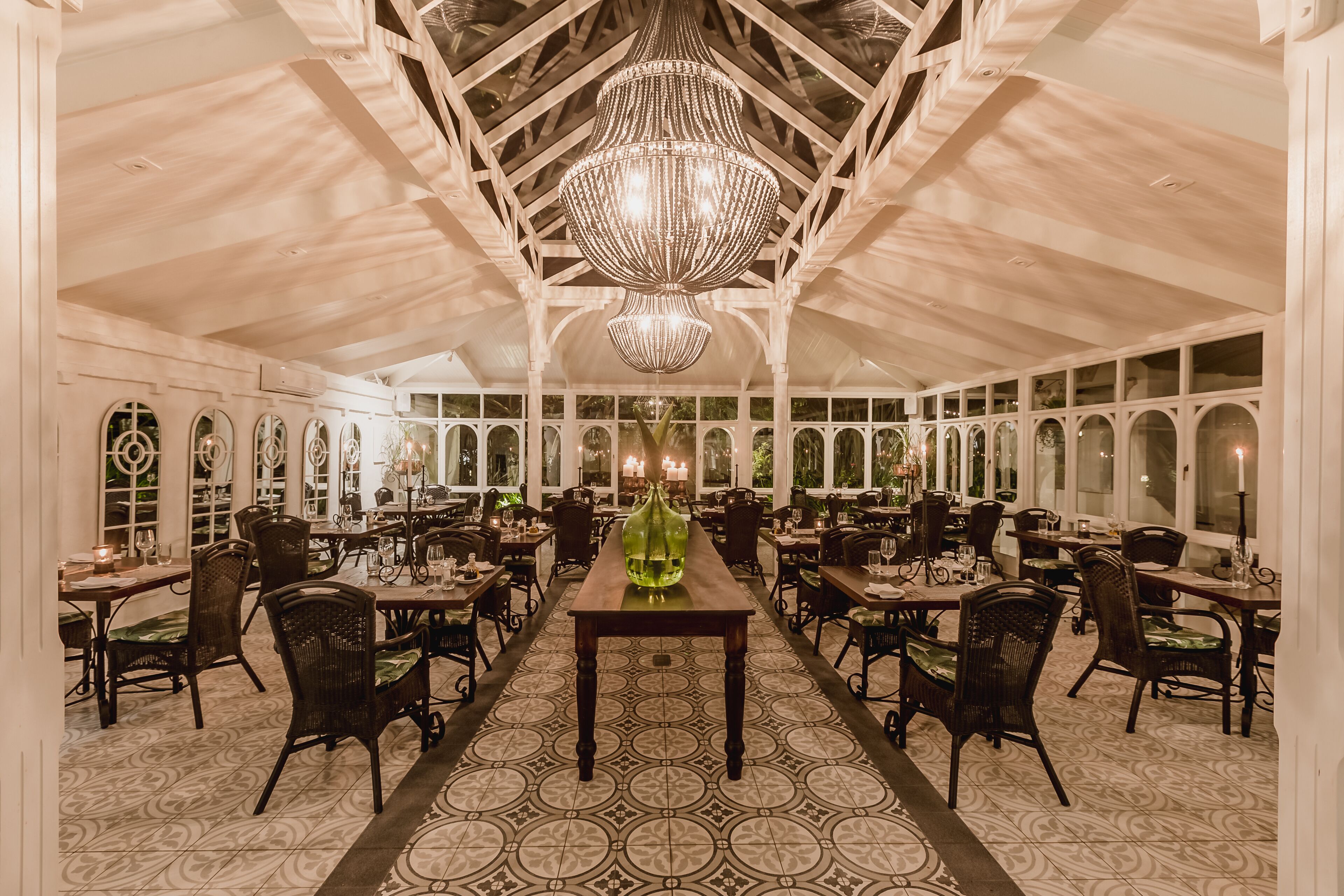 Une salle à manger spacieuse avec un plafond haut et un grand lustre. Les tables sont disposées avec des chaises, et une longue table au centre est agrémentée d’un vase vert.