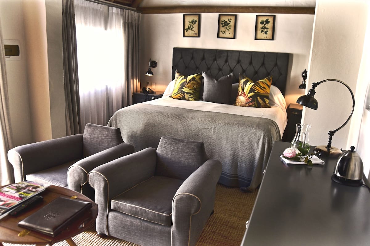 A hotel room with a large bed, grey armchairs, a bedside table with a lamp, and a vase with a flower.