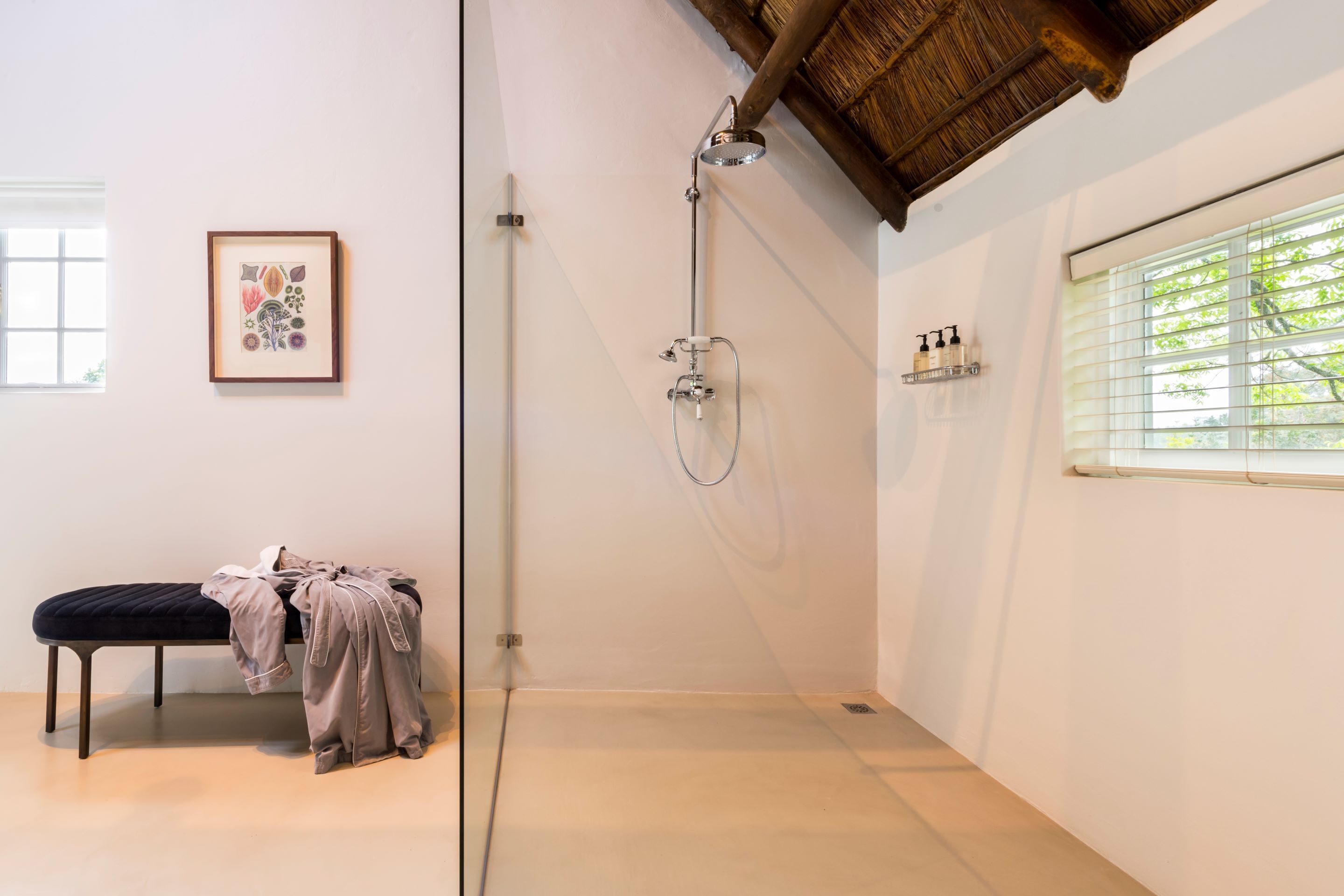A modern bathroom with a glass shower enclosure, a bench with clothes, a framed artwork, and a window with blinds.