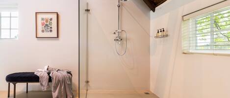 A modern bathroom with a glass shower enclosure, a bench with clothes, a framed artwork, and a window with blinds.