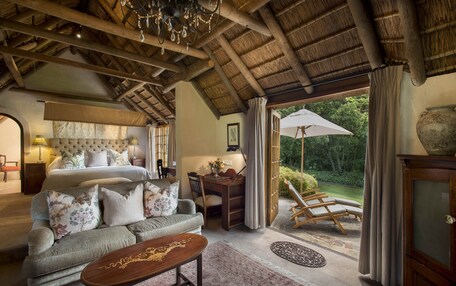 A spacious bedroom with a large bed, a sitting area with a sofa, a desk, and a view of the garden through a glass door.