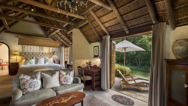 A spacious bedroom with a large bed, a sitting area with a sofa, a desk, and a view of the garden through a glass door. - Hunter's Country House (Plettenberg Bay)