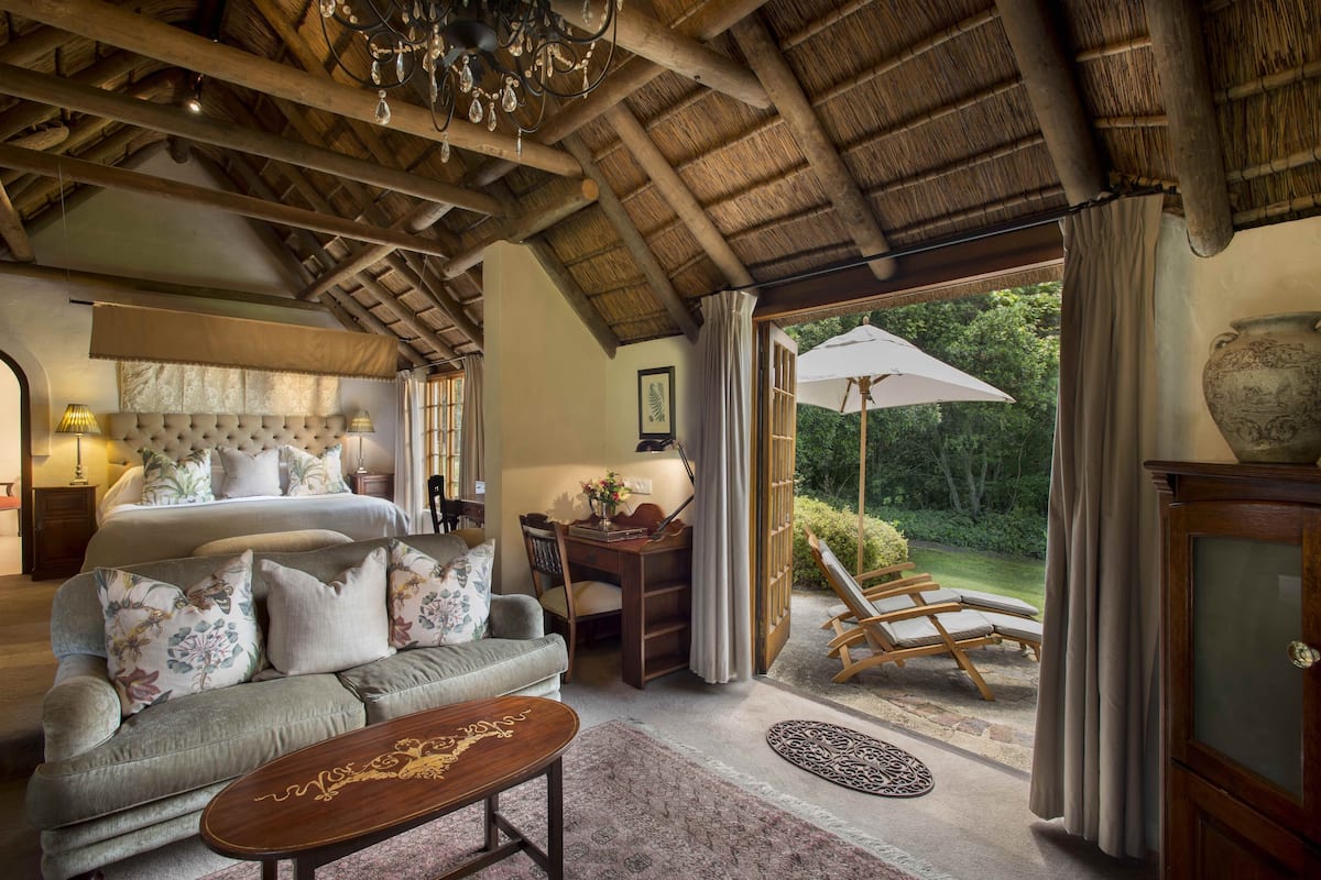 A spacious bedroom with a large bed, a sitting area with a sofa, a desk, and a view of the garden through a glass door.