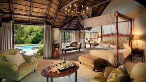 A spacious room with a four-poster bed, a sitting area with a sofa and armchairs, a dining table with a tray of items, and a view of a swimming pool through large windows. - Hunter's Country House (Plettenberg Bay)