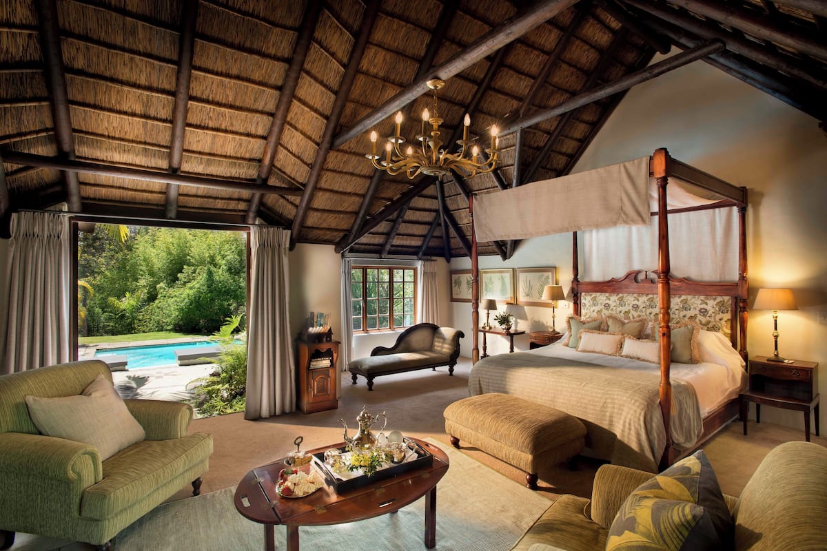 A spacious room with a four-poster bed, a sitting area with a sofa and armchairs, a dining table with a tray of items, and a view of a swimming pool through large windows.