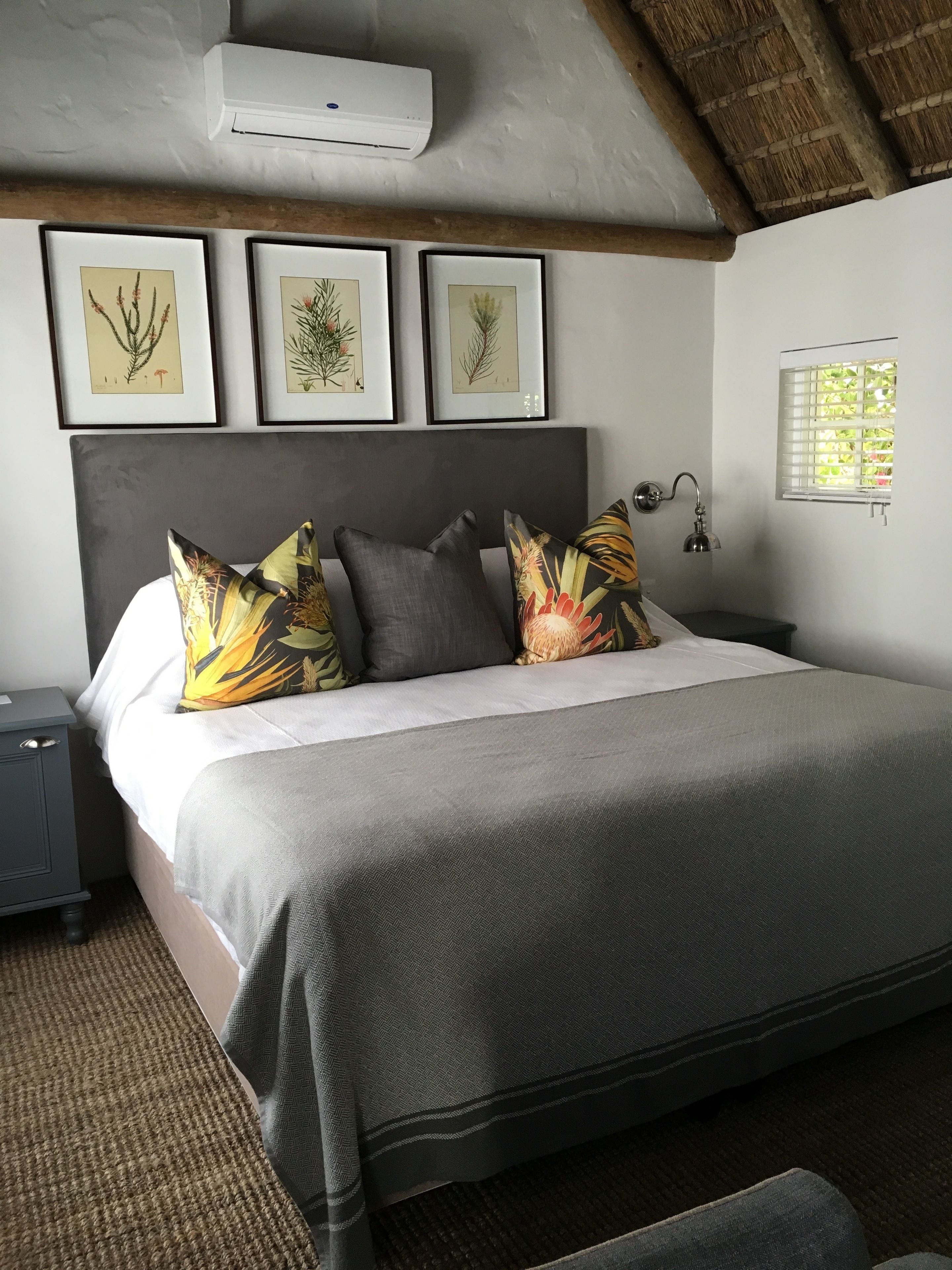Une chambre à coucher comprenant un lit, des oreillers, une table de chevet, un climatiseur fixé au mur, des illustrations botaniques encadrées et une fenêtre donnant sur un paysage verdoyant.