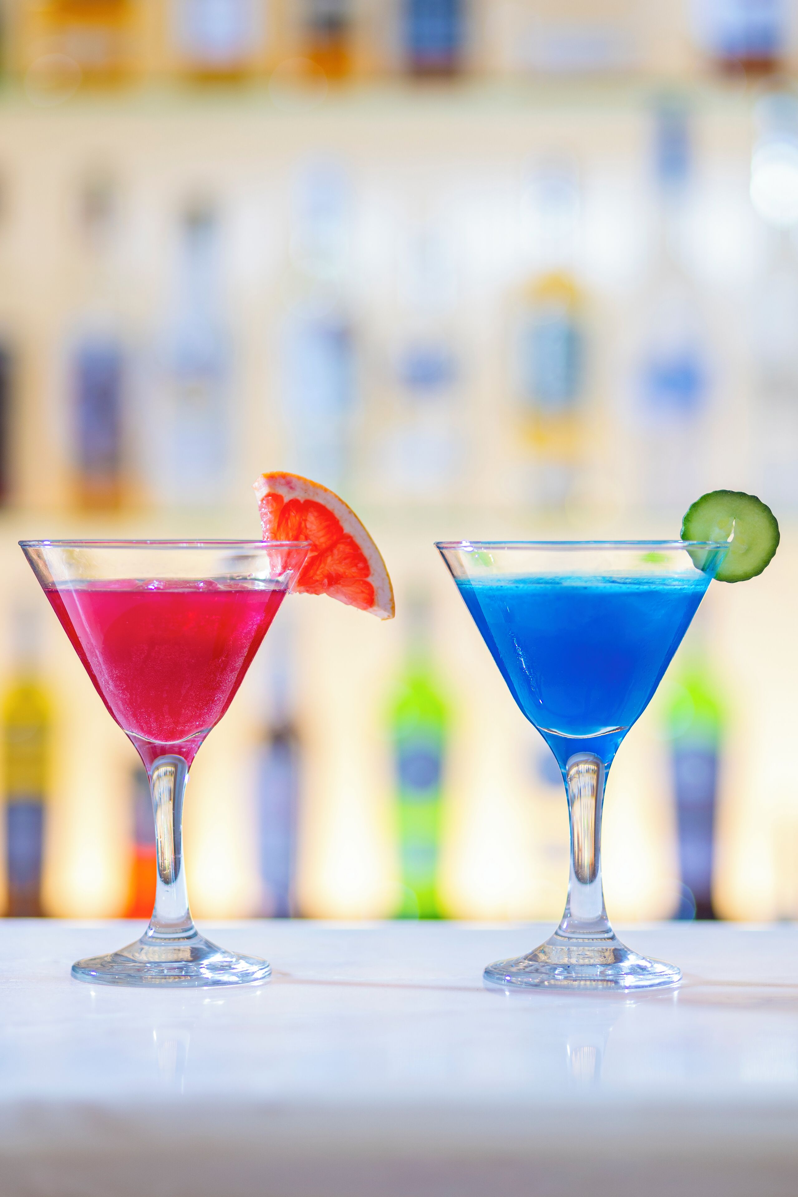 Dos copas de martini llenas de bebidas coloridas, una con una bebida roja y otra con una bebida azul, ambas adornadas con una rodaja de fruta.