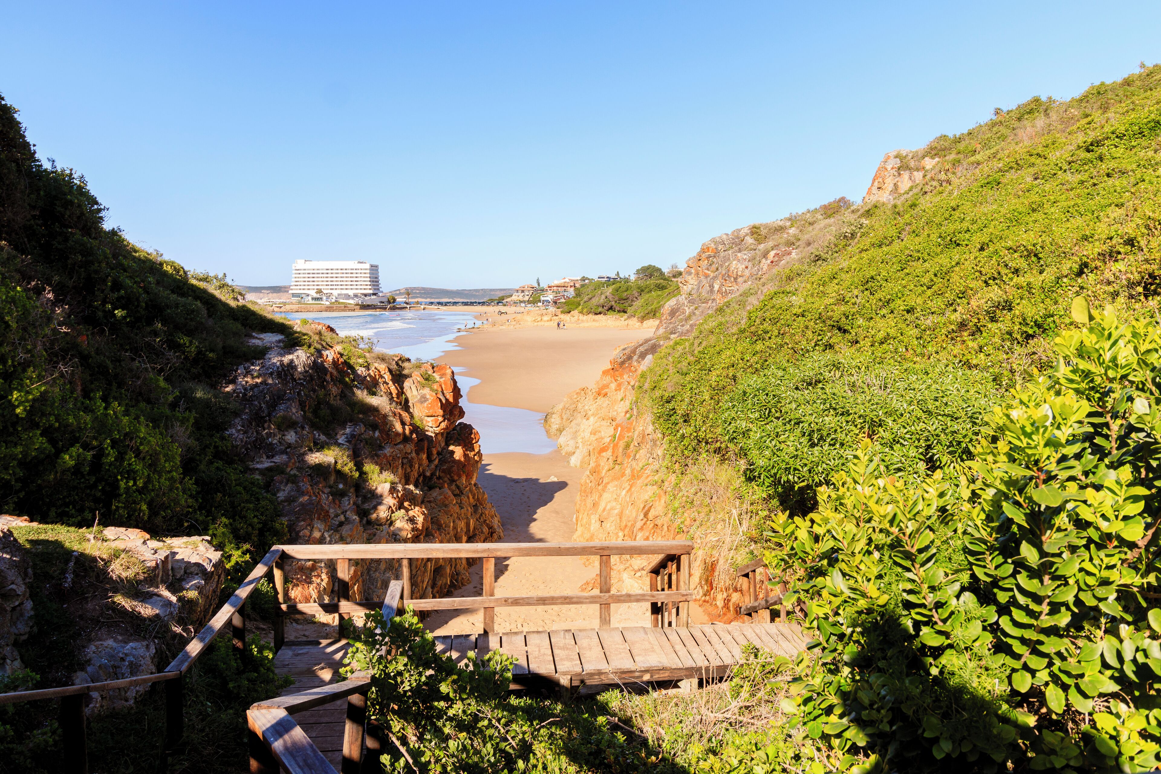 Un paseo marítimo que conduce a una playa con una terraza de madera y escaleras, rodeado de vegetación y un edificio a lo lejos.