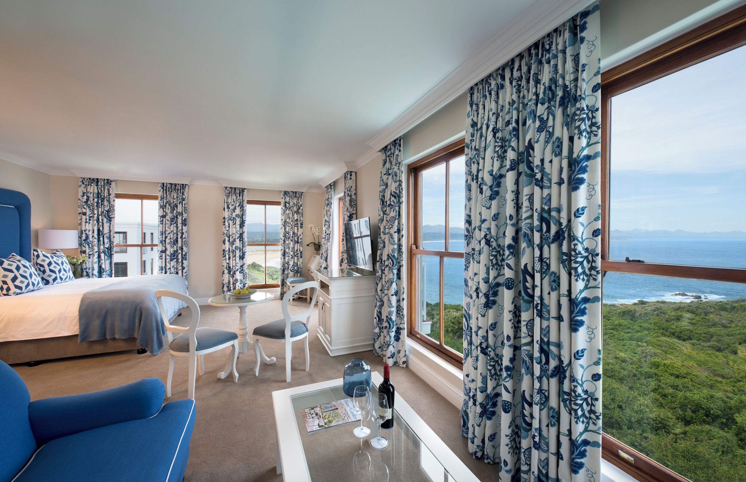 Une chambre d’hôtel avec un lit, un bureau, une chaise, une table avec une bouteille et des verres, et de grandes fenêtres donnant sur la mer.